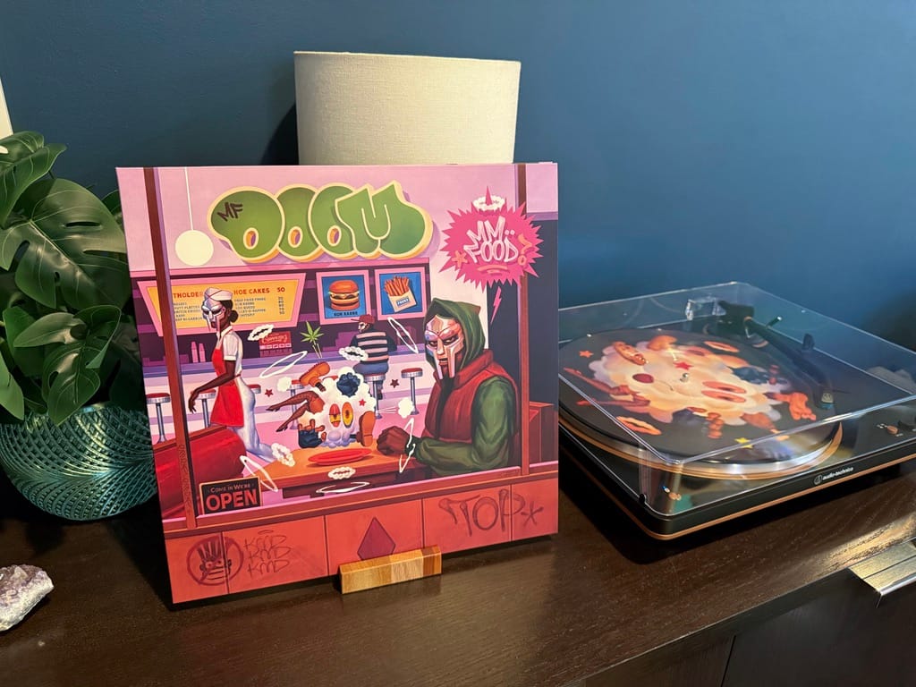 A vinyl record of MF DOOM's Mm..Food album playing on the turntable. The sleeve is propped up in front of a Monstera plant on a walnut cabinet. 